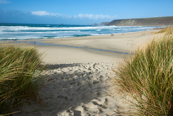 Sennen cove beach from dunes This landscape photograph depicts Sennen Cove beach in Cornwall, United Kingdom, as seen from the sandy dunes that frame the approach to the coast. The image was taken in the early afternoon during the autumn season, with bright sunlight casting clear shadows over the beach and grass-tufted dunes. The wide expanse of sand leads to the rolling waves of the blue sea, and the distant headlands typical of the rugged Cornish coast are visible on the horizon. The natural setting highlights the scenic beauty of Sennen Cove and its coastal surroundings, offering a clear view of the relationship between the beach, the Atlantic Ocean, and the characteristic landscape of Cornwall in the United Kingdom.
